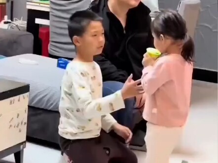 The little sister, having already had her yogurt, still wants to drink her big brother’s—just one se