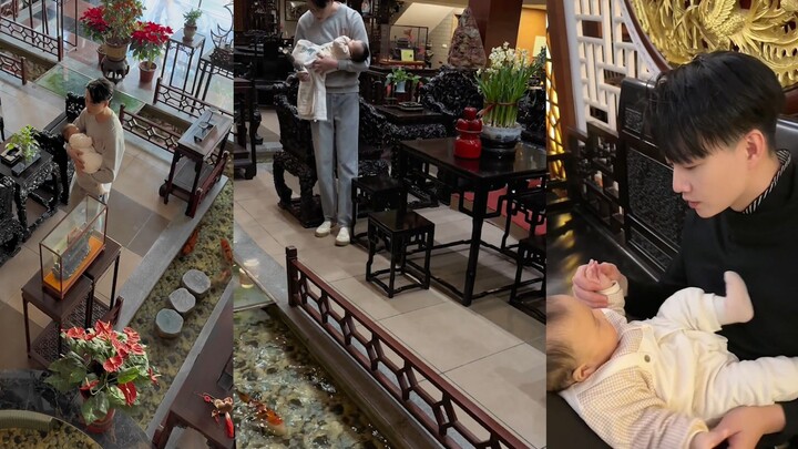 Người đàn ông bế con đi dạo trong nhà để dỗ ngủ, bước chân chậm rãi, nhẹ nhàng vỗ lưng bé; khung cản