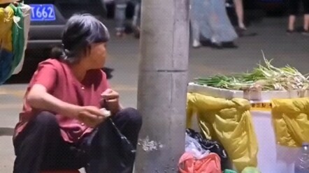 ผู้สูงอายุยังคงขายผักในยามค่ำคืน แล้วเหลียวมองเป็นระยะไปยังกลุ่มคนวัยเดียวกันที่กำลังเต้นรำอยู่ด้านห