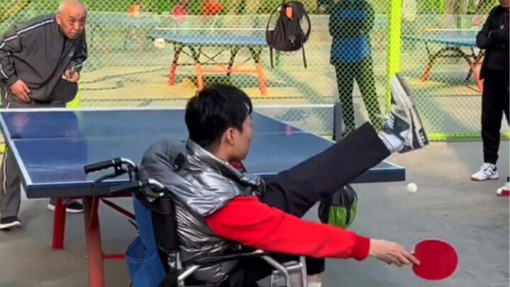 Di taman, seorang pria berada di kursi roda sambil bermain bola bersama seorang bapak; para penonton
