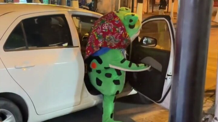 Encountered a frog mascot on its way home from work, hailing a taxi—its adorable struggle to squeeze