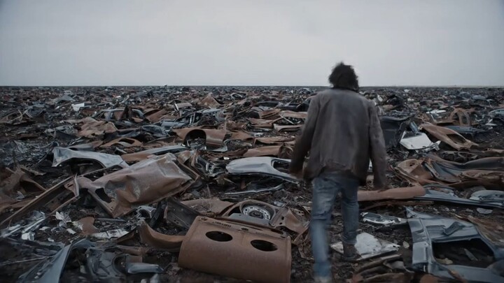 A man walks over a surreal realistic landscape where the ground is made only of car metal debris,